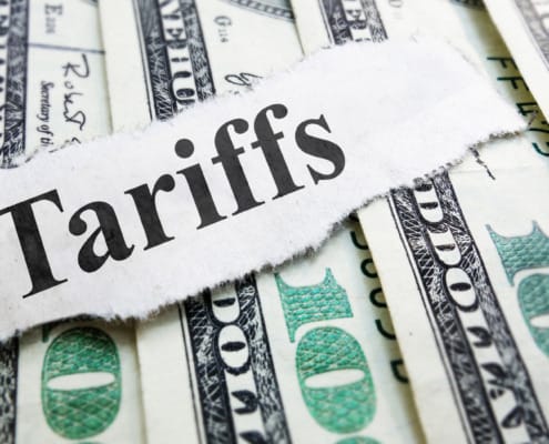 A close-up of a torn piece of paper with the word "Tariffs" written on it, placed on top of a pile of U.S. hundred-dollar bills, symbolizing the financial impact and economic implications of tariffs.