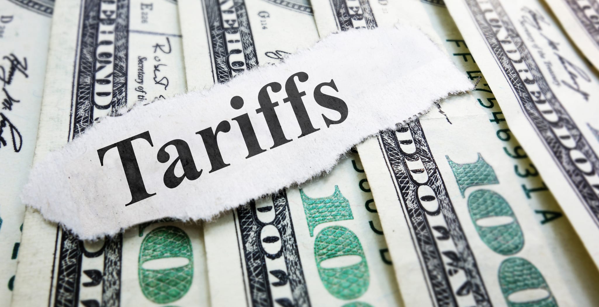 A close-up of a torn piece of paper with the word "Tariffs" written on it, placed on top of a pile of U.S. hundred-dollar bills, symbolizing the financial impact and economic implications of tariffs.