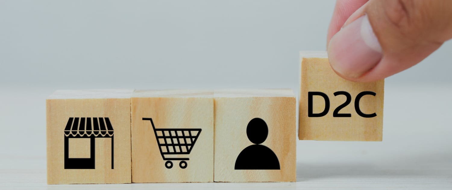 A hand placing a wooden block labeled "D2C" next to three other wooden blocks with icons representing a storefront, a shopping cart, and a person. The image symbolizes the direct-to-consumer (DTC) business model, illustrating the shift from traditional retail to direct sales channels that connect brands directly with consumers.
