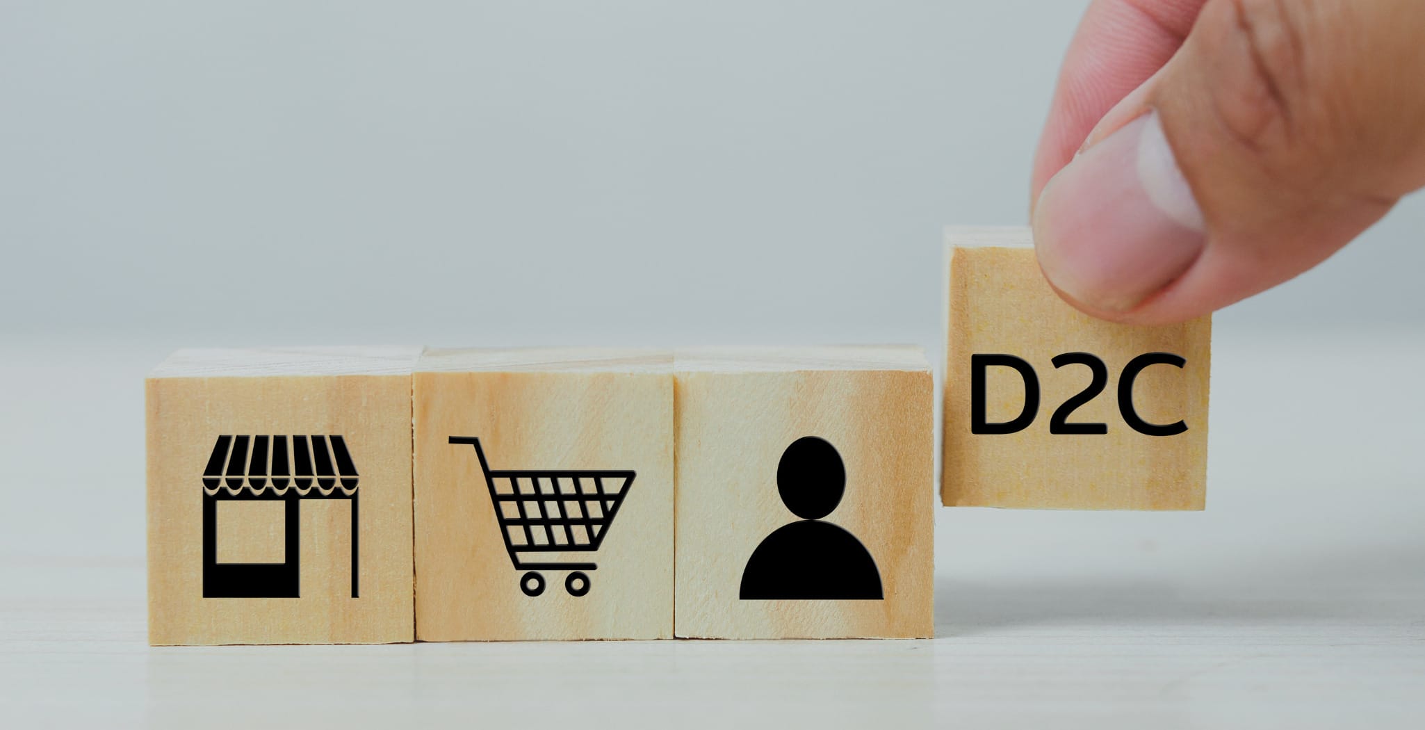 A hand placing a wooden block labeled "D2C" next to three other wooden blocks with icons representing a storefront, a shopping cart, and a person. The image symbolizes the direct-to-consumer (DTC) business model, illustrating the shift from traditional retail to direct sales channels that connect brands directly with consumers.