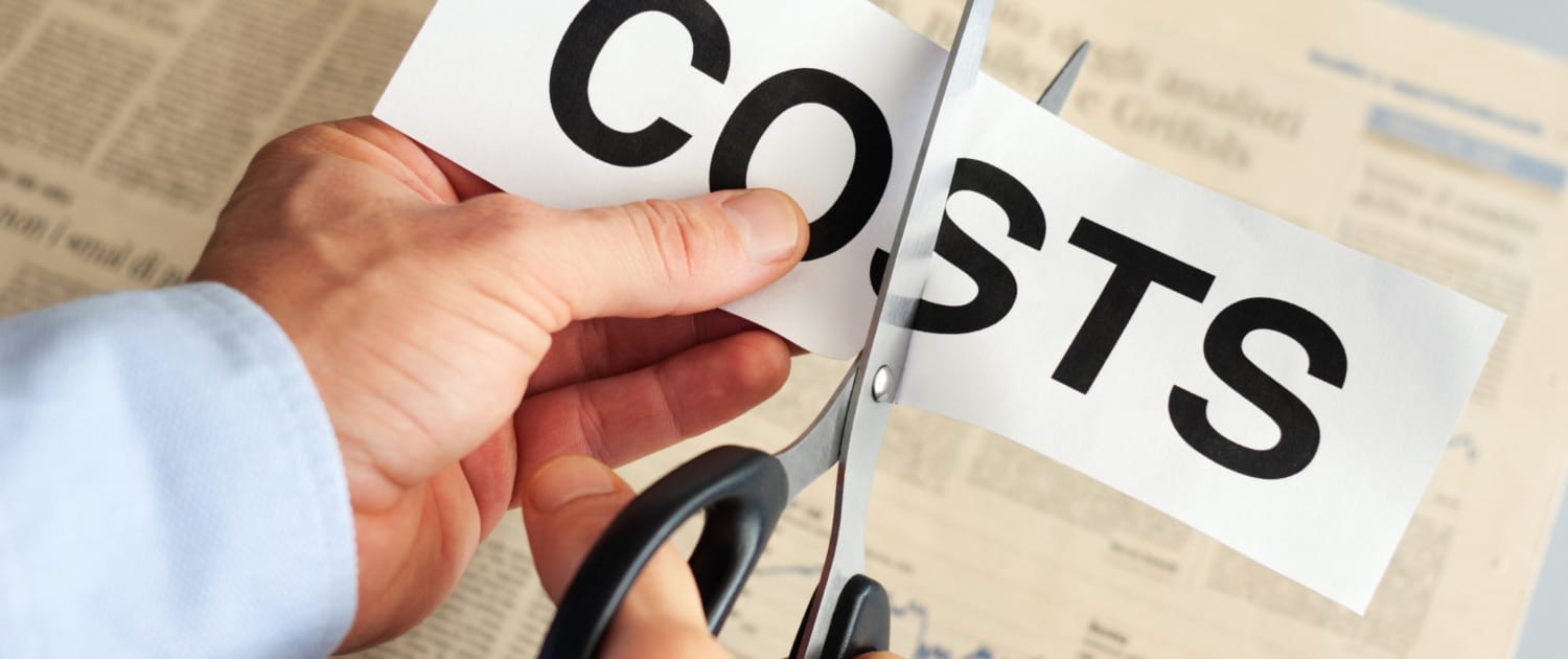 A close-up of a person in a blue dress shirt using scissors to cut a piece of paper with the word "COSTS" printed in bold black letters, symbolizing cost reduction. A financial newspaper with charts and articles is visible in the background.

