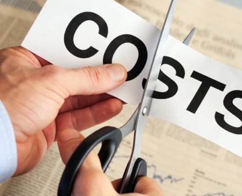 A close-up of a person in a blue dress shirt using scissors to cut a piece of paper with the word "COSTS" printed in bold black letters, symbolizing cost reduction. A financial newspaper with charts and articles is visible in the background.