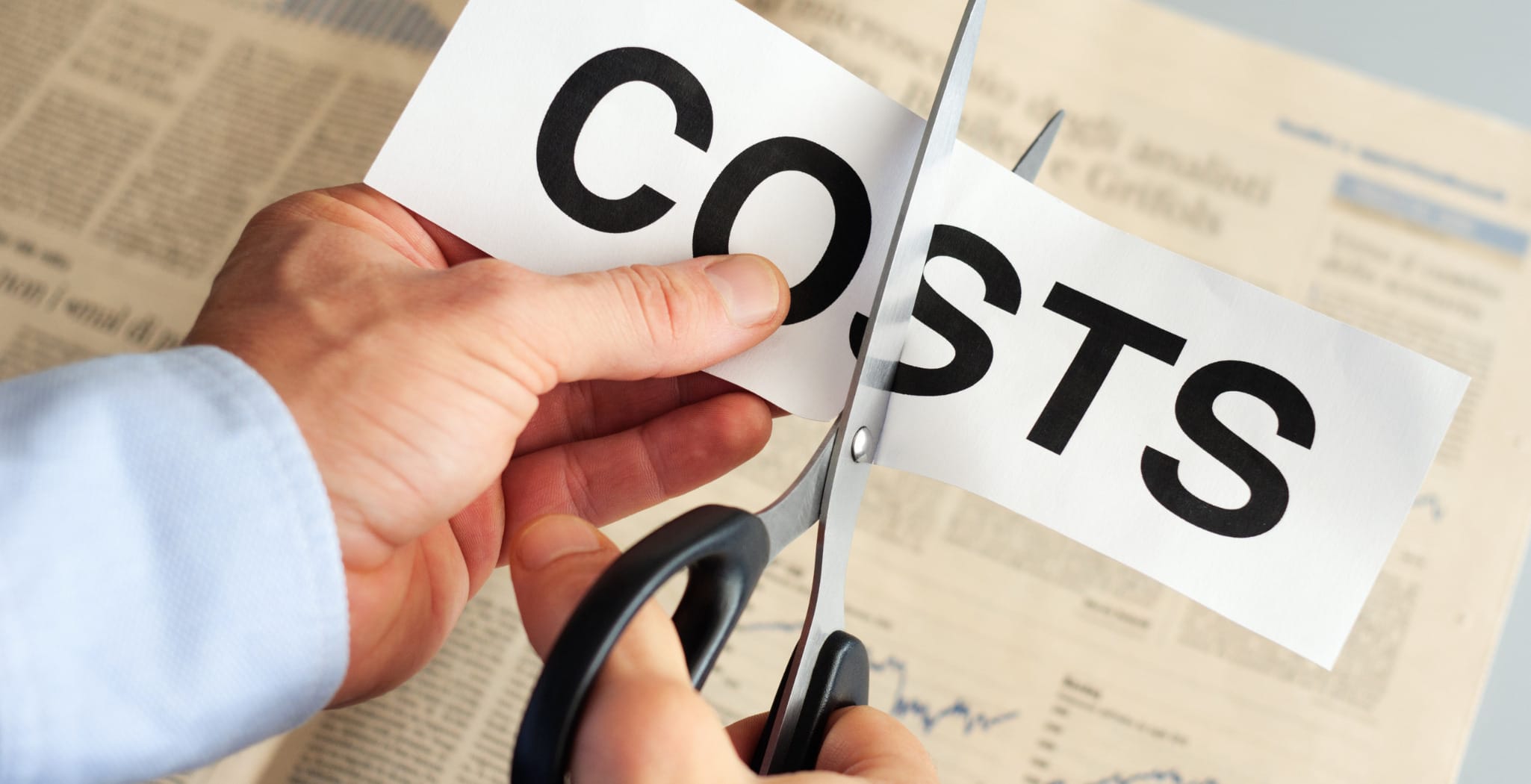 A close-up of a person in a blue dress shirt using scissors to cut a piece of paper with the word "COSTS" printed in bold black letters, symbolizing cost reduction. A financial newspaper with charts and articles is visible in the background.