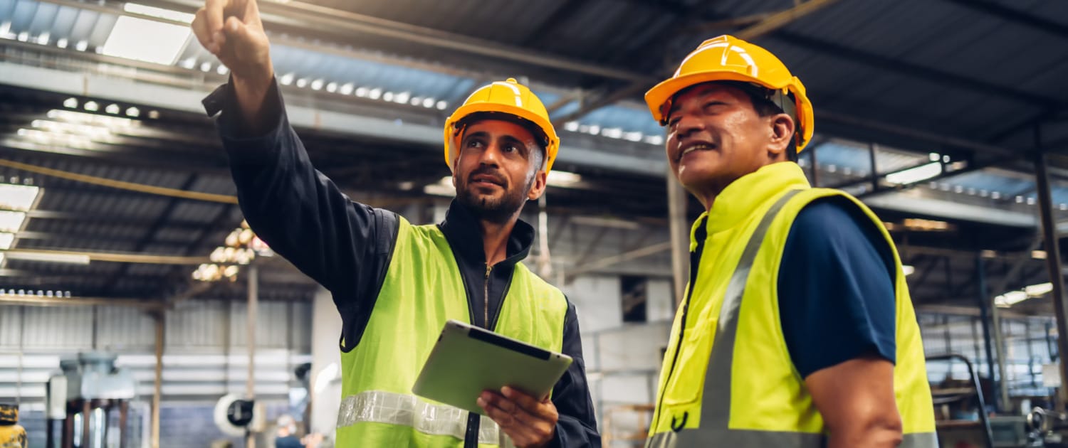 Two factory workers wearing yellow safety vests and hard hats stand inside an industrial facility. One worker, holding a tablet, gestures upward while explaining something to the other, who listens with a smile. Machinery and equipment are visible in the background, indicating a manufacturing or production environment.