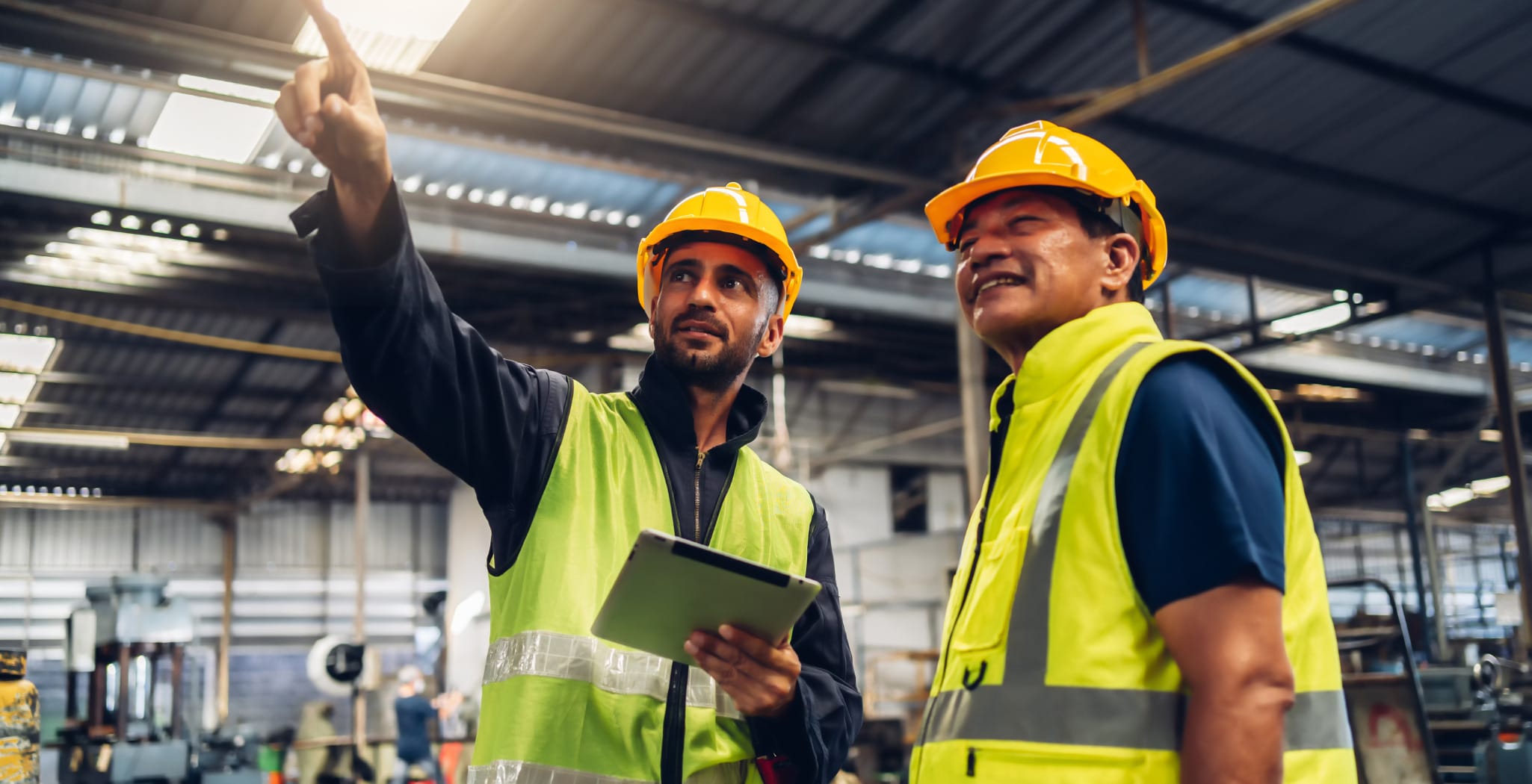 Two factory workers wearing yellow safety vests and hard hats stand inside an industrial facility. One worker, holding a tablet, gestures upward while explaining something to the other, who listens with a smile. Machinery and equipment are visible in the background, indicating a manufacturing or production environment.