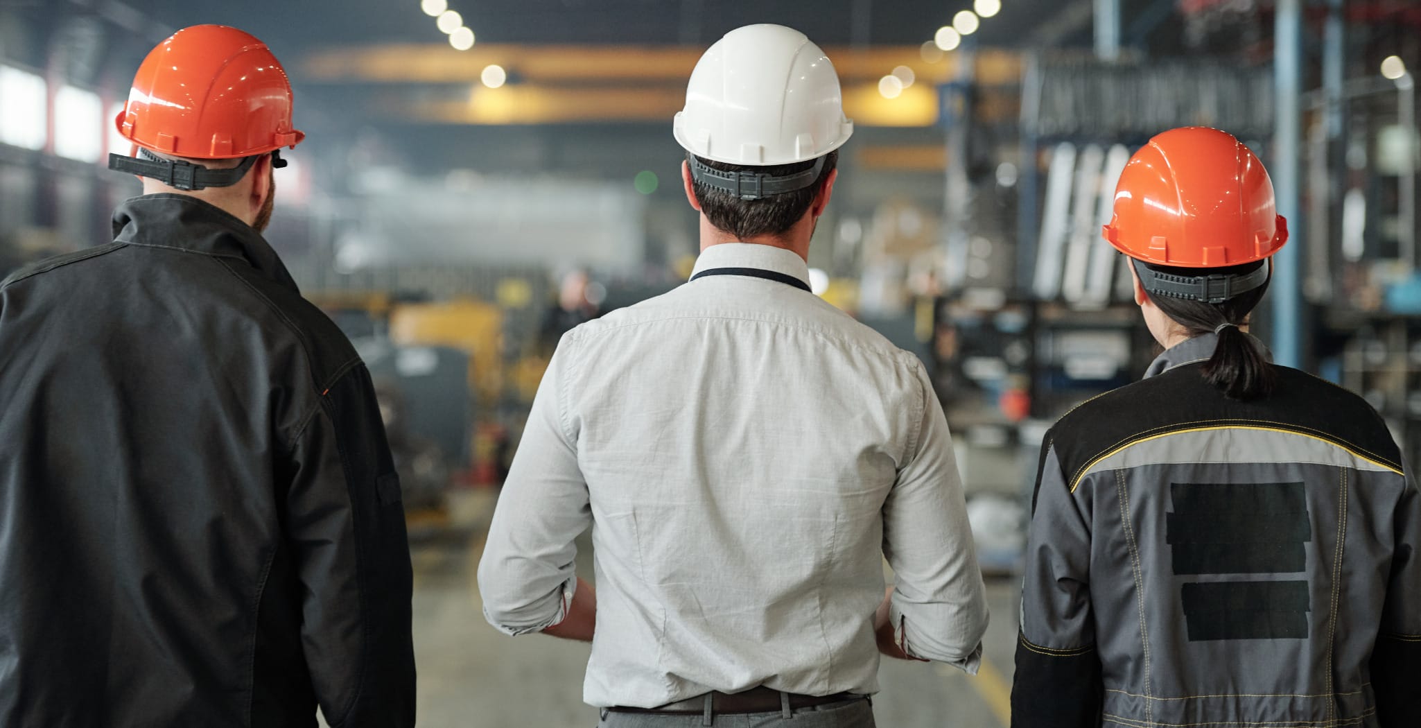 Three factory workers wearing safety helmets and work uniforms walk through an industrial facility, viewed from behind, symbolizing collaboration, manufacturing leadership, and production oversight.